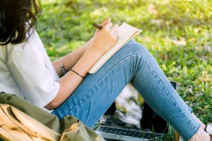 Woman with pen writing on a notebook sitting on grass in park